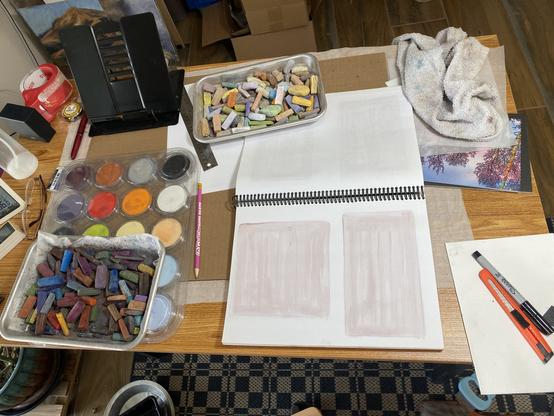 Small desk filled with stuff. Two trays of pastel sticks, pan pastel, and sketch book. Other junk.