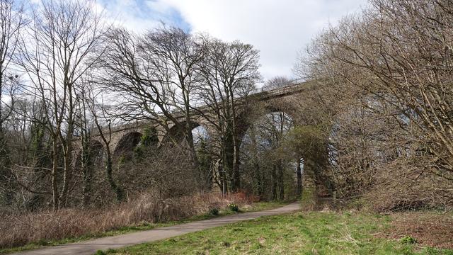 A railway viaduct (6 of the 7 arches visible) stretching across a valley with a path running underneath and trees bending in the wind