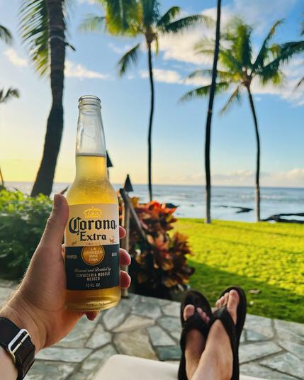 A Corona beer bottle in the foreground; flip-flopped feet, golden-hour grass, palm trees, and the Pacific Ocean in the background. A real shit view.