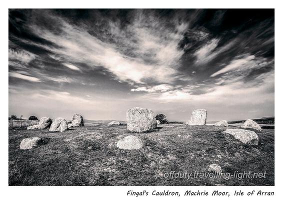 Toned black and white photo of a stone circle with a central stone, on grass, with clouds appearing to converge above the centre of the circle.