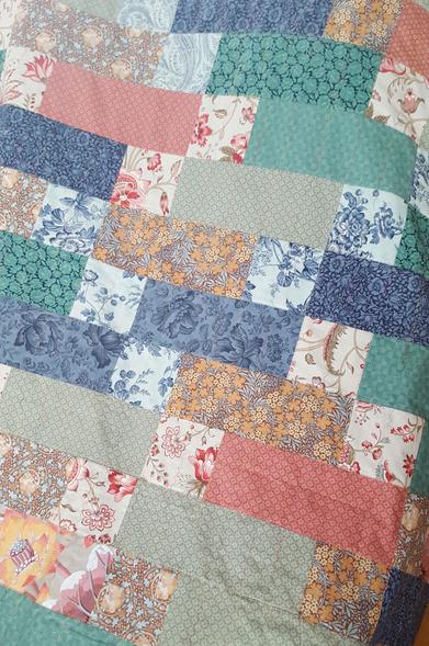 A closer up photo with the entire frame filled by the quilt. It is mostly random, but with pieces of it aligning in a geometric little wave of squares. The colours are greens, teals, blues and reds, with a little bit of brown, earth tones and the tiniest bit of yellow. Also gray! It looks pretty nice, though it has obviously not been ironed before this photo.