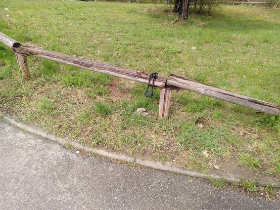 Grasfläche neben Asphaltweg, am Rand der Grasfläche ist ein niedriger baufälliger Zaun aus Holzstämmen. Über dem Zaun hängt ein Faltschloss.