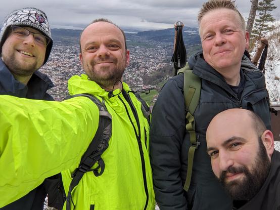 Gruppenfoto mit vier Männern im Vordergrund und dem Blick vom Berg auf den Bodensee im Hintergrund. Alle lächeln, einer hat eine leuchtende Jacke an.