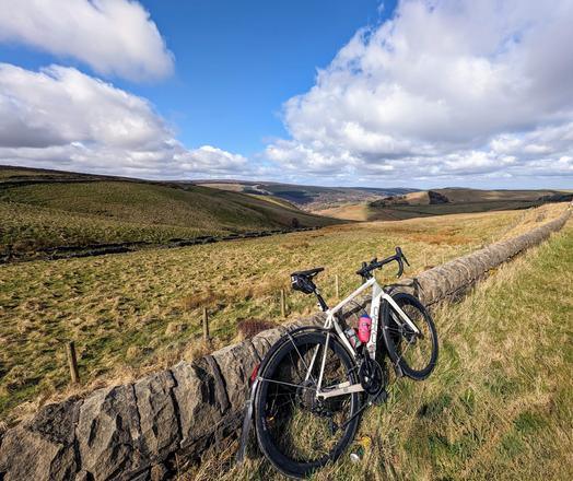 View from the top of Long Hill, Derbyshire. A white bicycle leaning against a low, stone wall. In the background are rolling grass moors topped with blue sky and white clouds.