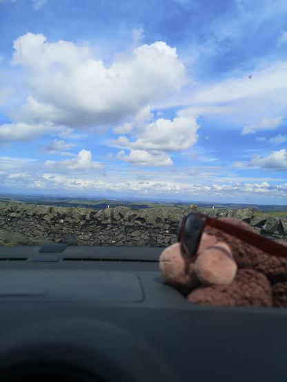 Vom Parkplatz an der Grenze an der A68 zwischen England und Schottland, kann man weit ins Land nach Schottland sehen. Der Parkplatz liegt auf der Spitze eines kleinen Berges. Vor uns sind Wiesen und weite Flächen im Sonnenlicht zu sehen. Darüber ziehen weiße Schäfchen Wolken ihren Weg am Himmel