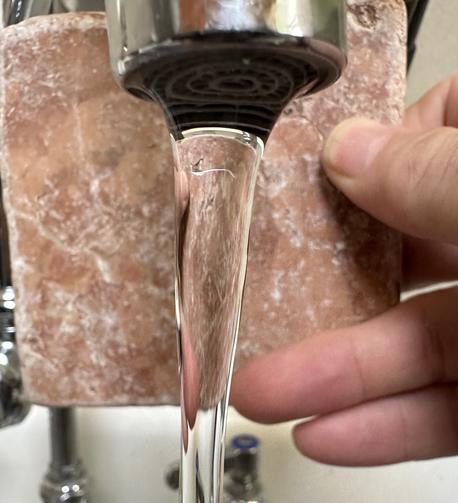 Closeup of laminar flow coming out of a faucet. A hand is holding a stone tile behind the flow to highly the crystalline clarity. The flow looks like a solid bar of glass, as if you could catch a hold of it and hang it on your yule tree.