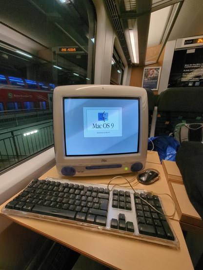 our friendly iMac G3, still laying on the same table as before. there's a keyboard and a mouse in front of it. the Mac displays a french Mac OS 9 splash screen
