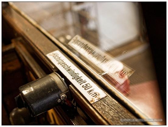 Weißes Emailleschild mit der schwarzen Aufschrift "Höchstgeschwindigkeit 50 km" innen auf dem verwitterten Holzrahmen der Windschutzscheibe eines historischen Schienenbusses aus den 1930er-Jahren; das Schild spiegelt sich in der Scheibe.