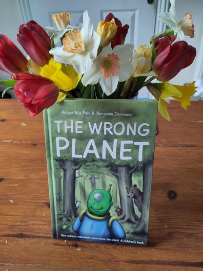 A book on a wooden table beneath a vase of yellow and white daffodils and red tulips. The book is THE WRONG PLANET by Holger Nils Pohl, illustrated by Benjamin Dammeier, subtitled: How autistic individuals experience the world. A children's book. The cover shows a green, one-eyed, smiling alien with an antenna on their head, wearing a blue and yellow space suit. The alien is surrounded by trees in a forest, with animals peeking out from behind the trees. There is a frog, a bear, an otter and a beaver, while two ducks are flying past.
