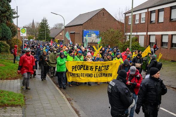 Fronttransparent des Demozugs für die Erhaltung Lützeraths. Auf einem Werbeplakat steht: „Wir bitten an Christi statt:“. Auf dem Front-Transpi steht: People & facts unite behind Lützerath“, Keyenberg, 12.01.2023