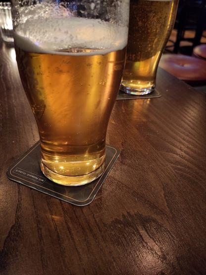 2 pint glasses of amber coloured beer on a brown pub table.