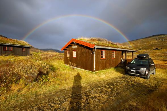 Intense rainbow over a wooden house in Norway. Our car is parked next to it. The sky is dramatically colored, with rain clouds and sunshine.