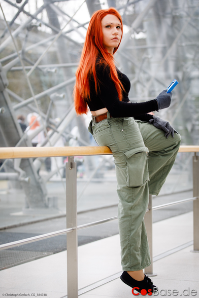 A cosplay of the character Kim Possible, sitting on a handrail. She is a girl with bright orange/red hair, wearing a black tank top, grey gloves, olive baggy pants and black sport shoes.