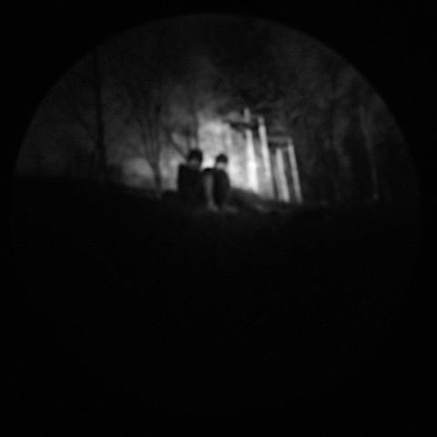 Ein pinhole-Bild. Zu sehen sind zwei Menschen, die vor Bäumen sitzen und in die Camera schauen. Sie haben dunkle Mützen auf, ihre Gesichter sind nicht zu erkennen. Das Bild ist schwarzweiß und wirkt sonderbar mystisch. Da die Leinwand im Inneren der Büchse rund ist, entsteht eine rundformatige Projektion.