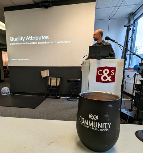 Cory Gwin speaking in front of a slide that says Quality Attributes, Define how well as system should perform some action, a quote from Michael Keeling's book Design It!