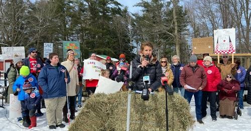 A woman speaking into a microphone behind a hay bale. Behind her is a crowd of people, many of them holding up signs that say, "Save Smiling Hill farm." There are people of all ages in the crowd, including children. Behind them is a pen with cows, and in the distance is a stand of tall pine trees.