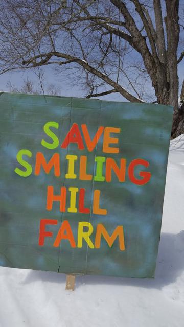 A sign stuck in the snow. In red, orange, green and yellow letters are the words, 
SAVE
SMILING
HILL
FARM.
The background of the sign is a combination of light blue and green spray paint.