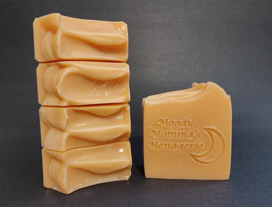 On the left are 4 bars of pale orange soap stacked on their side to show the top detail. The bar on the right in stamped with "Moody Momma's Menagerie" and a crescent moon