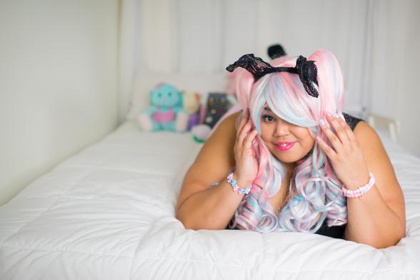 An African American woman who wears plus size clothing lies on a white canopy bed in a pink and black dress, black lace bunny ear headband, candy-colored plastic jewelry, and cotton candy-colored blue and pink wig. A variety of stuffed animals is collected at the head of the bed behind her.