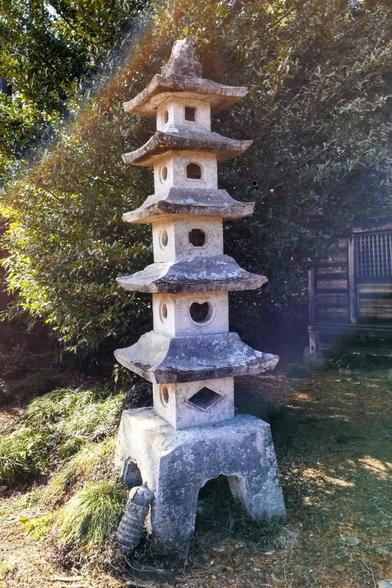 A multi tiered stone lantern. Each segment is a box made of stone with a roof that extends a third out in all four directions. There are openings in the sides. Each segment is not that dissimilar from a bird house. The bottom one has a footprint of about a foot (or 30cm) squared. It sits on what I would describe as a kindergarten stool and each lantern segment up gets about 10 per cent smaller. There are five of them and the top one's roof is steeper and bigger. The sun is coming in from the side and my camera recorded a slight lens flare in the top third of the image.