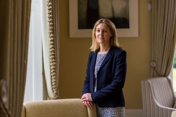 A photograph of a woman in a royal blue blazer over a blue and white polka dot dress. She is stood with her hands resting on a large armchair in a room with large windows and brown drapes that match the chair