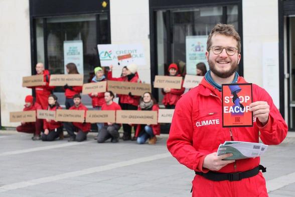Action devant une agence Crédit Agricole pour dénoncer le projet Eacop. Un militant en combinaison rouge tient un tract Stop EACOP face au photographe. Au second plan, l'agence Crédit Agricole, avec des militants et militantes devant des pancartes dénonçant les discours de la banque à ce sujet.