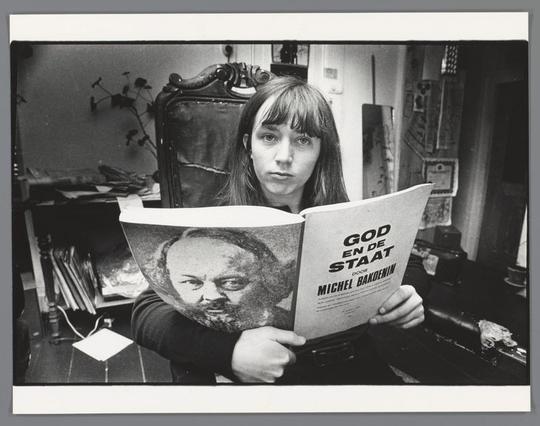 This is a black and white photograph featuring an individual holding a book. The person appears to be engaged in reading or studying, as suggested by the focused gaze on the book cover. The book is titled "God and the State," authored by Michel Bakoenin, and has a subtitle indicating it's the "Greek" edition. It mentions that it includes a foreword by Jean-Paul Sartre.

The person is seated indoors, with a room containing various items visible in the background. The setting seems to be casual and personal, possibly a living space or a study area.

In the bottom right corner of the image, there's text that provides some context for the photograph: "Hillie Molenaar met het boek 'God en de staat', door Michel Bakoenin, in haar huis aan de Hoogte Kadijk, Amsterdam, Ed van der Elsken, 1969 - 1971." This text translates to "Hillie Molenaar with the book 'God and the State' by Michel Bakoenin, at her house on the High Road in Amsterdam, Ed van der Elsken, 1969-1971."

The style of the image is vintage, and it conveys a sense of nostalgia. The photograph has a timeless quality to it, capturing a moment from nearly five decades ago.