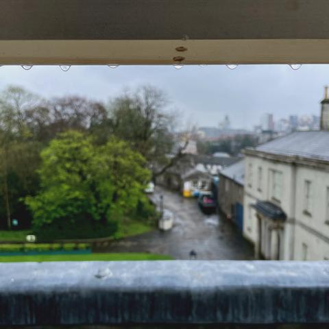 Raindrops hanging from a window frame with a blurred view of a street and buildings in the background.