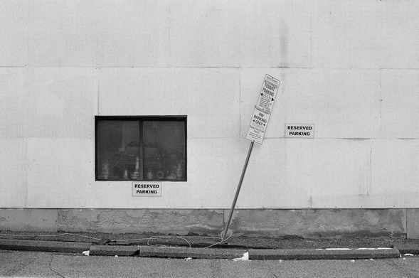 A black and white photograph of a wall bordering a parking lot. Signs state: “reserved parking”, “authorized tenant parking”, and “pay parking only” in apparent contradiction of each other.