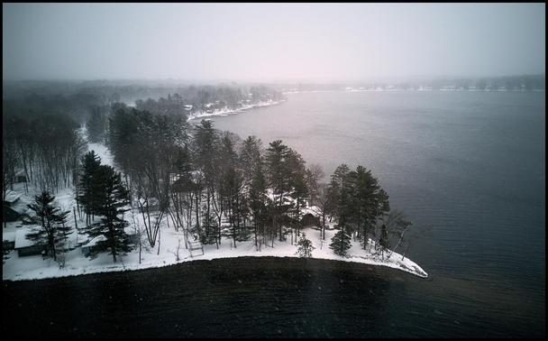 Aerial photo taken just about the tree line along the shores of an open lake, during a snow storm. The ground and cabins are all covered in snow. The sky is dark and snow can be seen in the air.