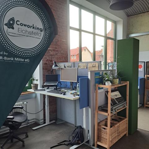 Blick auf einen Arbeitsplatz mit Stehtisch und Bildschirm Laptop 

Links steht eine kleine Flagge in grün und Logo des coworking eichsfeld