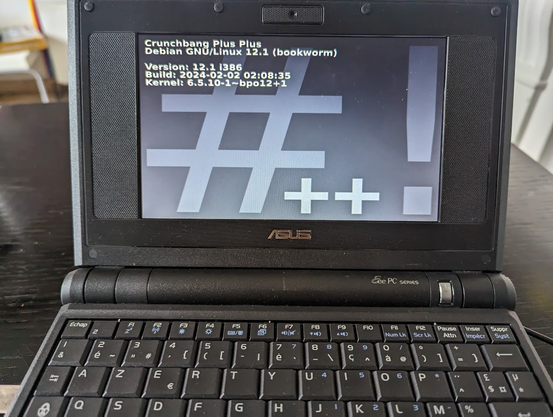 Photo d'un Asus EEE PC 701 de 2009, en couleur noire, posé sur un bureau noir lui aussi, avec l'écran allumé qui montre la page d'accueil de la distribution CrunchBang Plus Plus.