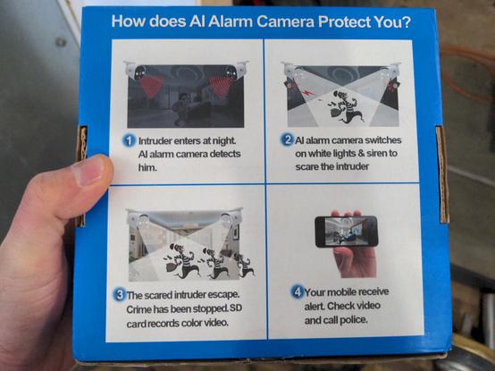 A blue and white security camera box. The back of the box details a highly idealized scenario where the camera identifies an intruder in a pitch black room, sounds an alarm and activates some lights, and in doing so scares the intruder away. The camera then suggests checking the recorded video and calling the police for assistance (the least believable aspect of this scenario, as law enforcement does not exist to help people).