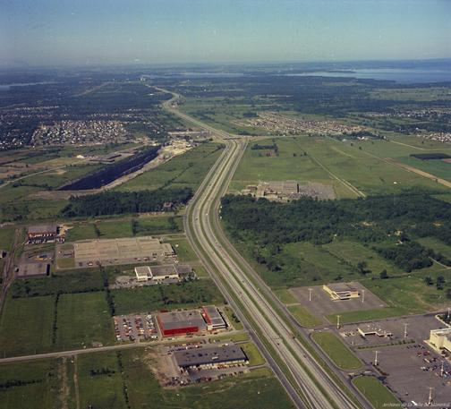 Aerial photo of western portion of the Island of Montréal.  Mostly green because the image is from the 1970s.  Light colored divided highway runs bottom to top into the distance (Autoroute 40, part of Trans-Canada Hwy).    Some industries along the highway. Large stone quarry in upper left.  Edge of a shopping mall (Centre Fairview) in lower right)