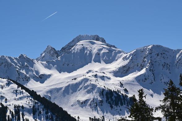 Foto von verschneiten Bergen vor wolkenlosem blauen Himmel in dem man ein Flugzeug mit Kondensstreifen sieht.

In Bildmitte sieht man eine Sesselbahn und daneben von SkifahrerInnen total verspurte Hänge.