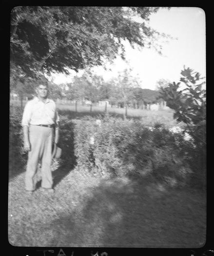 The image is a black and white photograph. It features a person, presumably male, standing in what appears to be an outdoor setting, likely a backyard or garden. He is wearing a button-up shirt and slacks, with his hands clasped in front of him. In the background, there are trees and what seems to be a fence. The photo has a vintage appearance, suggesting it may have been taken some time ago. There are no texts visible in the image.