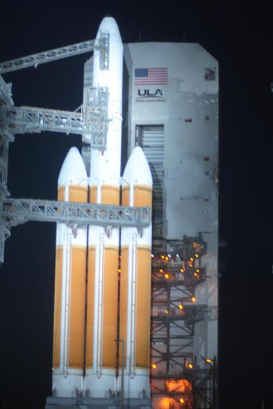 An outdoor, night time photograph of a rocket on its launch pad. The rocket has a central cylinder that has orange foam on the bottom and white above. Two shorter cylinders are attached to the central orange core and are orange themselves except for white nose cones.
There is a white hanger in the background, and truss connections to the rocket from the right side of the image.