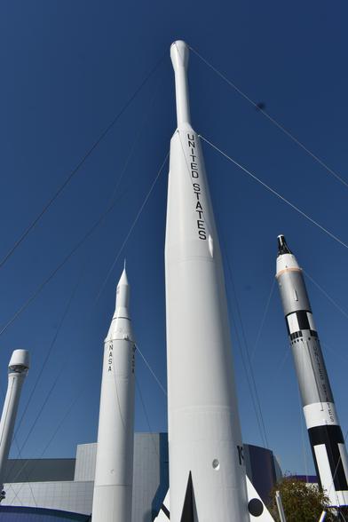An outdoor, daytime photograph of a white rocket against a blue sky with other rockets in the background, all held in place by wires.
The central rocket is white and is a cylinder with a narrowing top, which turns into a narrow cylinder that has a large "bud" on the top.
Behind it are two other rockets, one white and one gray and white and black.
The center rocket has the words "UNITED STATES" written vertically in black letters.