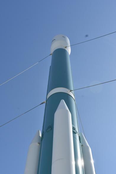 An outdoor photograph of a teal colored rocket against the clear blue sky.
The rocket is a cylinder with teal pain, and has three smaller cylinders with nose cones attacked around it. Above the teal section is a white cone-like shape that holds the payload.