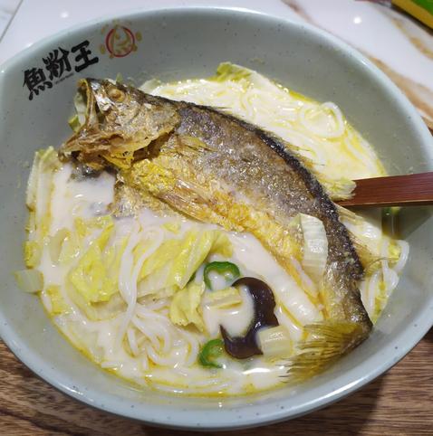 A bowl of milky broth is filled with white rice noodles, wood ear, and napa cabbage. All of it is topped by a whole fish – a yellow croaker that has been lightly pan-fried.