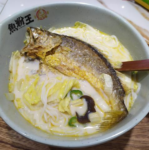 A bowl of milky broth is filled with white rice noodles, wood ear, and napa cabbage. All of it is topped by a whole fish – a yellow croaker that has been lightly pan-fried.