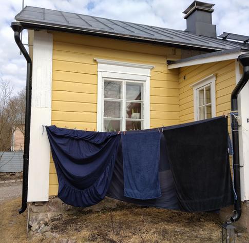 Tummat lakanat ja pyyhkeet kuivuvat ulkona narulla taustallaan keltainen vanha hirsitalo.