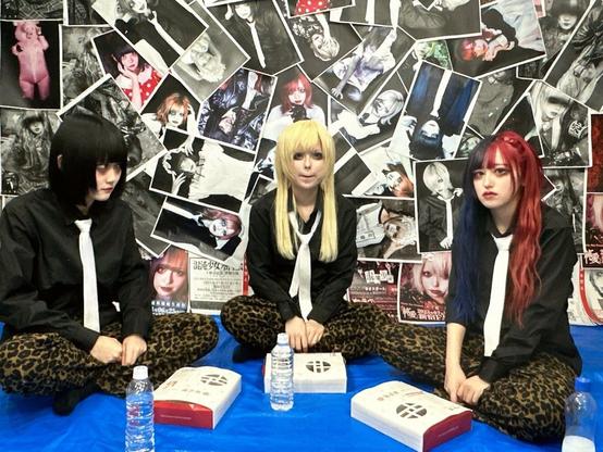 The three sit around with closed bento's in front of them. Behind them is a wall of pictures.