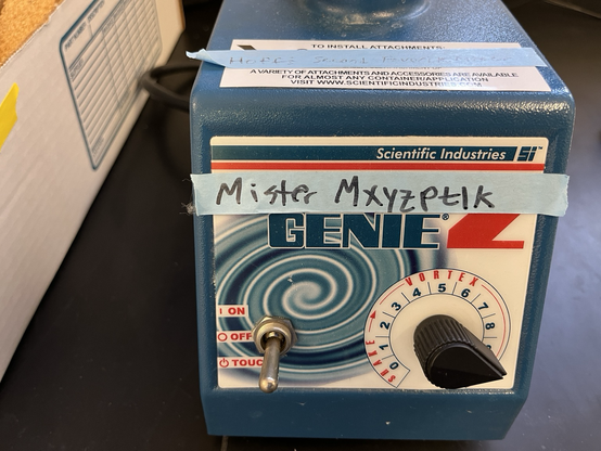 Machine for swirling test tubes to mix them (Vortex Genie 2: a blue box with a toggle switch and speed dial on the front and a cupped moving unit on top) with an pale blue tape label on which “Mister Mxyzptlk” is written in black sharpie. Sitting on a black epoxy resin lab bench with lab detritus scattered nearby