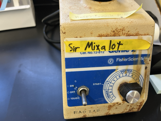 Machine for swirling test tubes to mix them (old Vortex Genie 2: a corroded tan box with a toggle switch and speed dial on the front and a cupped moving unit on top) with a yellow tape label on which “Sir Mixalot” is written in black sharpie. Sitting on a black epoxy resin lab bench with lab detritus scattered nearby