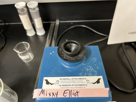 Machine for swirling test tubes to mix them (Vortex Genie 2: a blue box with a toggle switch and speed dial on the front and a cupped moving unit on top) with a pale pink tape label on which “Mixxy Elliot” is written in black sharpie. Sitting on a black epoxy resin lab bench with lab detritus scattered nearby