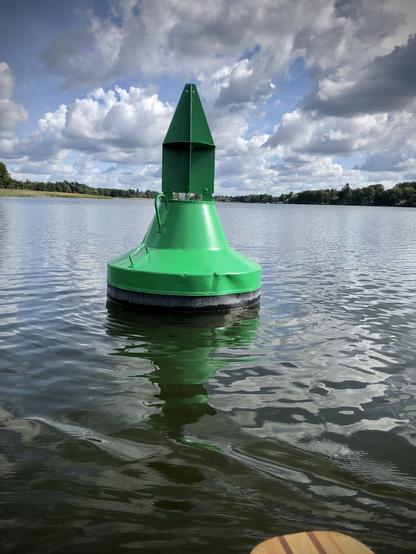 Eine grüne Boje, die auf dem Wasser schwimmt, mit einer bewaldeten Uferlinie und einem bewölkten Himmel im Hintergrund.