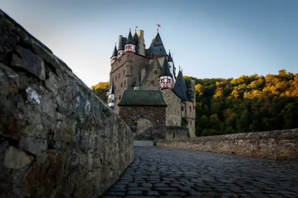 A photograph of Eltz Castle, taken from the access to the outer gate. Next to the photographer is a wall. The ground is paved with cobblestones. Behind the gate, the castle rises into the blue morning sky. To the right is the forest with its autumn leaves shining orange in the sunrise.