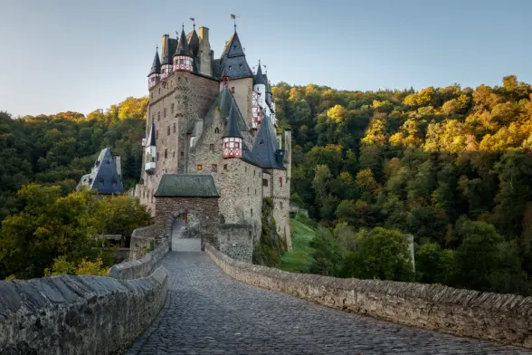 A photograph of Eltz Castle, taken from the access to the outer gate. The ground is paved with cobblestones. Behind the gate, the castle rises into the blue morning sky. To the right is the forest with its autumn leaves shining orange in the sunrise.