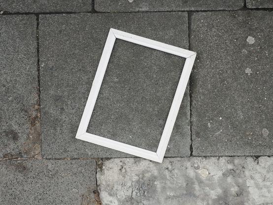 Von oben: Ein leerer, weißer Bilderrahmen liegt auf grauem Gehwegpflaster